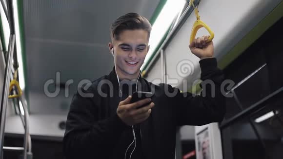 戴着耳机的迷人男人的肖像抓住扶手在公共交通工具上看智能手机上的视频城市视频的预览图