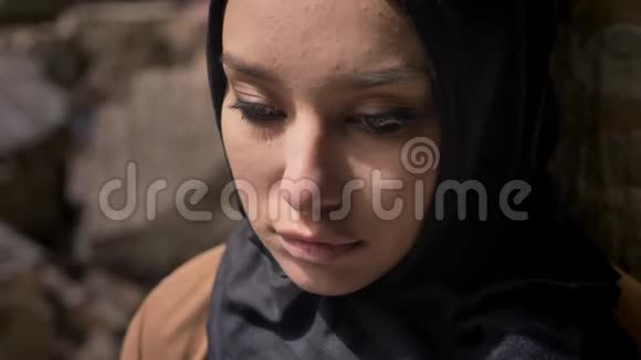 穆斯林年轻女子戴着黑色头巾站在砖墙旁边看着摄像机美丽迷人的眼睛视频的预览图
