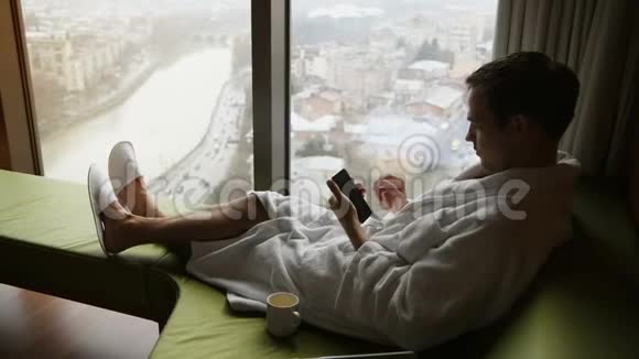 年轻人在智能手机上发短信坐在靠近窗户的公寓的床上有很好的城市景观视频的预览图
