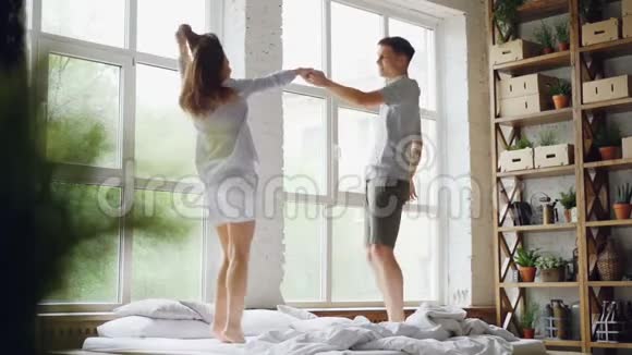 慢动作可爱的情侣在床上跳舞在周末的早晨在阳光下一起跳跃和大笑视频的预览图