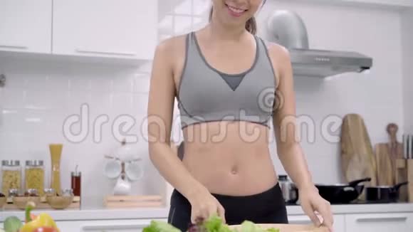年轻的亚洲女人感到快乐的舞蹈听音乐在厨房里做沙拉在运动服装中有很多营养视频的预览图