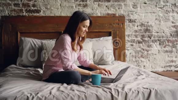穿着休闲服装在笔记本电脑上打字坐在阁楼风格的床上微笑的年轻女子动作缓慢视频的预览图