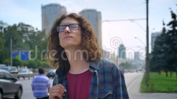 年轻的书呆子戴着眼镜留着长长的卷发等待出租车或公共汽车向前看站在附近的街道上视频的预览图