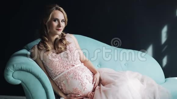 美丽的年轻孕妇坐在沙发上看着相机双手放在肚子上慢慢视频的预览图