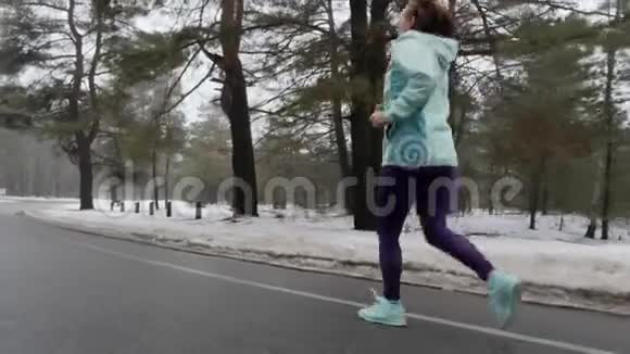 年长的白种人女人戴着耳机在冬天的雪地公园里跑步后面跟着射击慢动作视频的预览图
