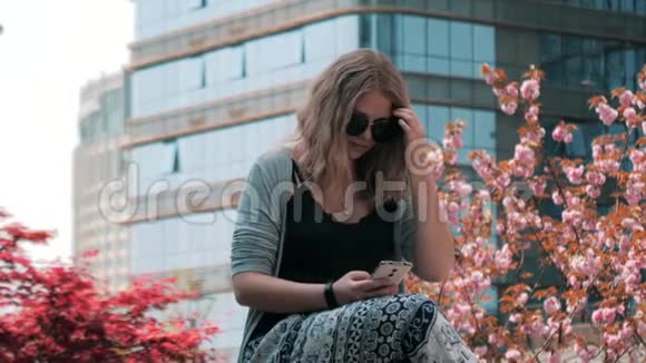 一位年轻漂亮的欧洲女孩戴着太阳镜坐在街上用智能手机连接摩天大楼视频的预览图