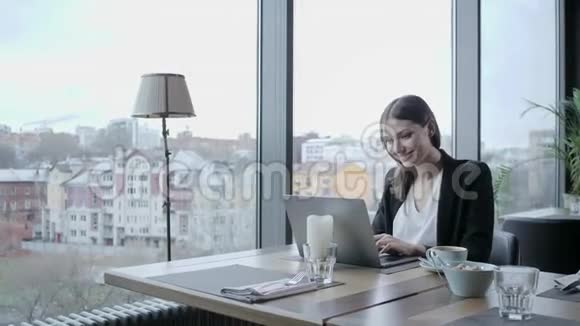 年轻女子笑着坐在木桌旁的咖啡店里桌子上是灰色铝制笔记本电脑女生博客浏览视频的预览图