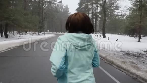 年长的白种人女人戴着耳机在冬天的雪地公园里跑步后面跟着射击慢动作视频的预览图