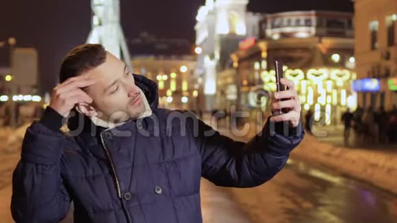 年轻的大胡子男在城市背景的夜晚拍搞笑自拍视频的预览图
