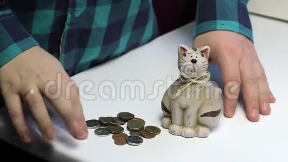 桌子上是一只猫形的储蓄罐一个人从桌子上捡起硬币把它们放在储蓄罐里视频的预览图