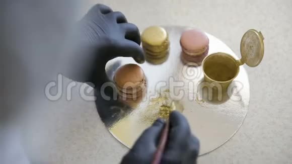 女人戴着黑色手套用刷子在厨房里用金色的水花装饰粉红色的马卡龙视频的预览图