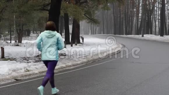 年长的白种人女人戴着耳机在冬天的雪地公园里跑步后退静态镜头视频的预览图