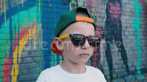 一个5岁的男孩戴着帽子戴着太阳镜在墙边的相机上涂鸦一个戴着太阳镜的孩子视频的预览图