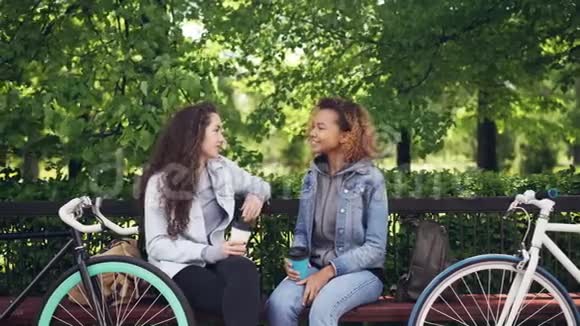 富有魅力的年轻女性白人和非裔美国人坐在长凳上聊天喝着外卖咖啡视频的预览图