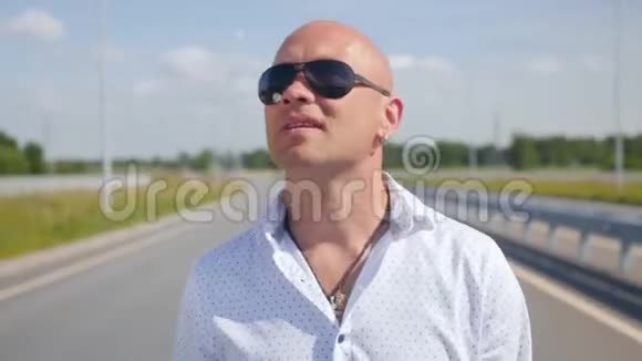 一个戴太阳镜的秃头男人在路上唱歌和散步的肖像中弹视频的预览图
