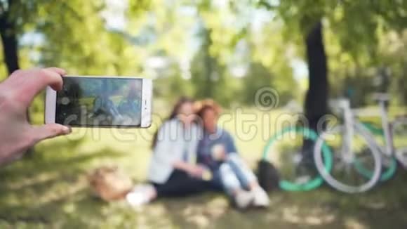 朋友正在拍摄两个漂亮女孩坐在公园的草坪上用手拿着手机专注于智能手机视频的预览图