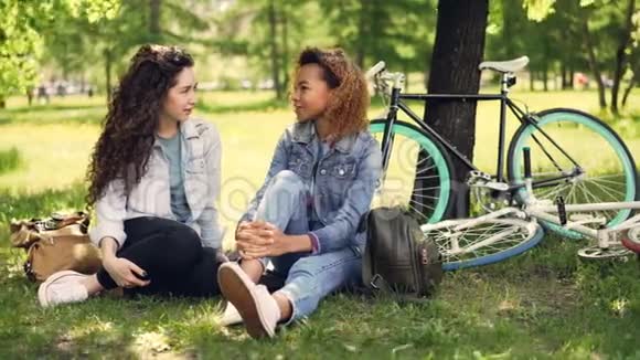 非裔美国女孩正和她的白人朋友在公园的草坪上交谈现代自行车和背包是可见的视频的预览图