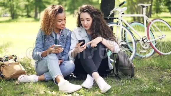 穿牛仔服装的漂亮女孩正在使用智能手机看屏幕坐在公园里谈笑风生视频的预览图