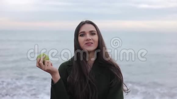 日出时一个穿暖和外套的漂亮女孩在沙滩上嚼一个绿苹果的特写镜头视频的预览图