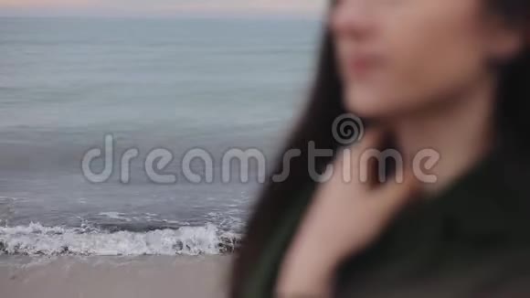 一个年轻女孩站在凉爽的春天的海滩上手抚摸着她的脖子和一件深绿色的外套视频的预览图