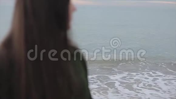 海边一个穿着深绿色外套的长发女孩的近距离模糊的侧视对平静大海的关注重点视频的预览图
