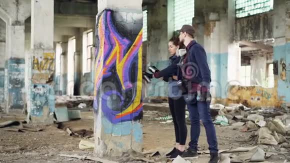 城市画家正在向有魅力的年轻女子解释如何涂鸦她们站在柱子旁边被损坏了视频的预览图