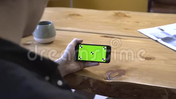 坐在木桌旁看着手机的彩色钥匙绿色屏幕一个穿着黑色衬衫的年轻人的肩膀上的景色视频的预览图