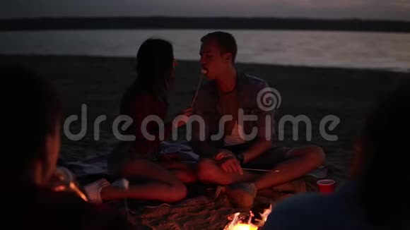 漂亮的白种人夫妇用棉花糖在棍子上互相喂食在篝火前接吻朋友都挂了视频的预览图