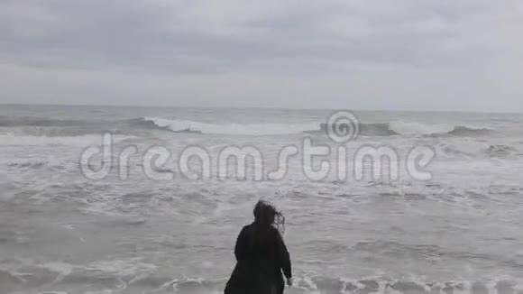 一个美丽的女孩在寒冷的一天自由地跑到波浪状的大海抚摸着水跑回来视频的预览图