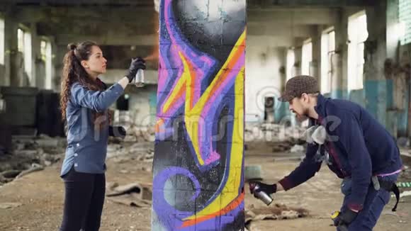 两位有技术的涂鸦艺术家留着胡须的家伙和迷人的年轻女子在废弃的仓库里一起工作视频的预览图