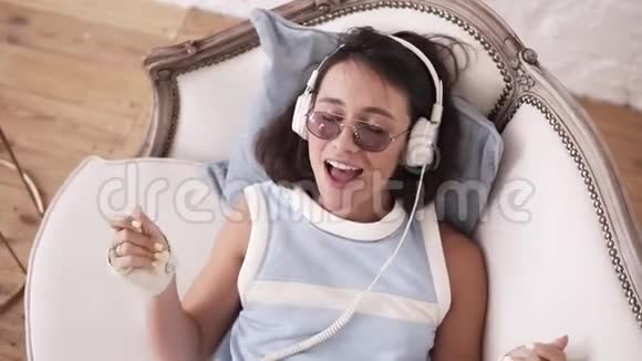 20的时髦女孩戴着耳机眼镜和朋克皮自行车手躺在沙发上唱歌视频的预览图