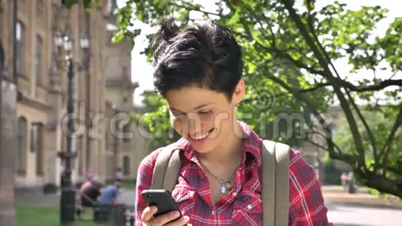 快乐美丽的女人留着黑色短发打着电话对着镜头微笑站在大学附近的街道上视频的预览图