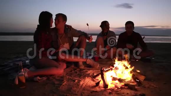 两对年轻夫妇坐在篝火旁的海滩上在木棍上烤肉吃一起度过快乐时光视频的预览图