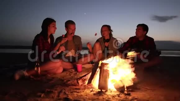 朋友们坐在篝火旁的格子布上用木棍煎食物吃东西的加速镜头视频的预览图