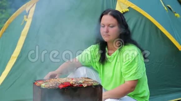 一个年轻的女人坐在一个帐篷背景的森林里在烤架上准备烧烤中间计划视频的预览图