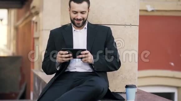 年轻的大胡子商人在平板电脑上看新闻休息时在办公楼附近喝咖啡视频的预览图