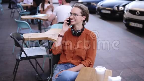 快乐自信的金发女人穿着牛仔布和橙色衬衫坐在街头咖啡馆的椅子上她用智能手机说话视频的预览图