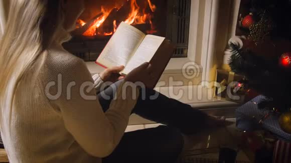 4k视频年轻女子在圣诞树旁夜间燃烧的壁炉旁读书喝茶视频的预览图