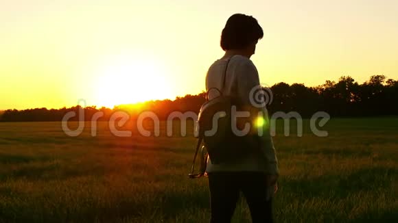 一个女孩在夕阳背景下背着背包在绿色草坪上行走的缓慢动作视频的预览图