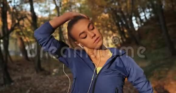 在公园里工作穿着蓝色夹克的年轻女子准备在美丽的秋天公园里跑步伸展脖子视频的预览图