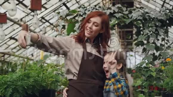 漂亮的温室女主人和她可爱的女儿正在果园里用智能手机自拍摆姿势和拍照视频的预览图