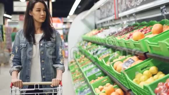 漂亮的年轻女子沿着水果和蔬菜排走推着购物车看着有机食品视频的预览图