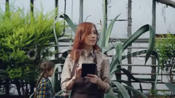 年轻的女农民正在温室里散步检查植物和使用平板电脑而她的女儿则在看着大片视频的预览图
