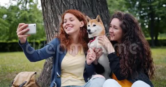 快乐的女孩朋友在公园里和可爱的狗一起用智能手机摄像头自拍视频的预览图