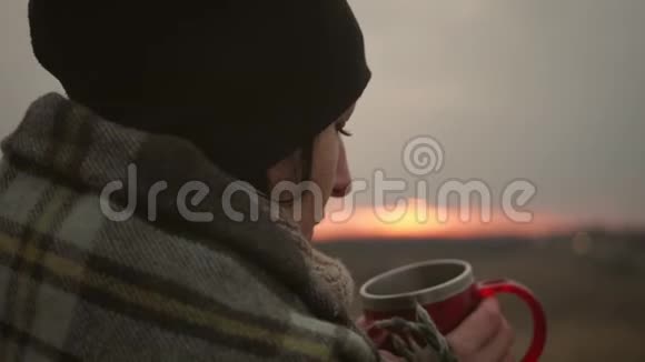 年轻的旅行女孩裹着格子在日落时用一杯饮料放松孤独自由孤独的概念视频的预览图