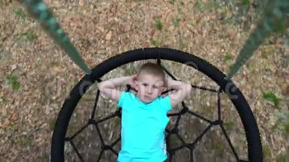 这位穿着蓝色t恤的蓝眼睛五岁男孩躺在一个圆秋千上双手放在脑后看着视频的预览图