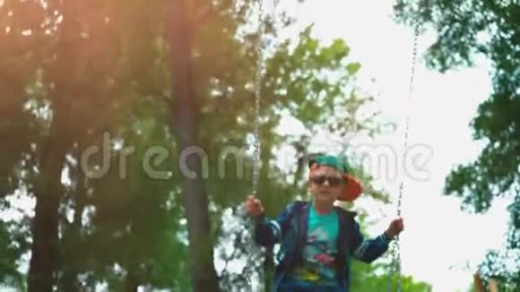 小男孩戴着太阳镜戴着绿色帽子在秋千上荡秋千一个5岁的孩子在孩子们秋千上玩得很开心视频的预览图
