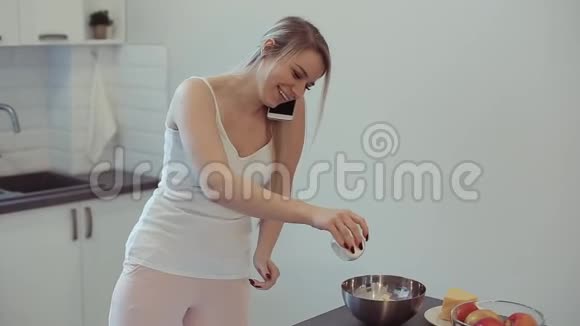 美丽的白种人女孩穿着白色衬衫在轻盈的厨房里做饭在手机上说话微笑着视频的预览图