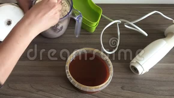 一个女人取下搅拌机的盖子用勺子混合切碎的花生和种子来制作卤汁容器旁边是视频的预览图
