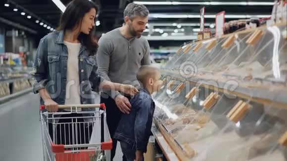 母亲父亲和儿子正在超市里买食物指着架子上的塑料盒子说话视频的预览图